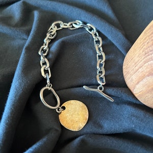 May include: A silver chain bracelet with a large, round, gold-toned pendant. The bracelet has a toggle clasp.