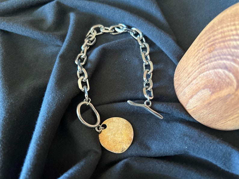 May include: A silver chain bracelet with a large, round, gold-toned pendant. The bracelet has a toggle clasp.