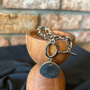 May include: A silver chain necklace with a large, round pendant. The chain is made of thick, linked metal and the pendant is smooth and shiny. The necklace is displayed on a wooden stand.