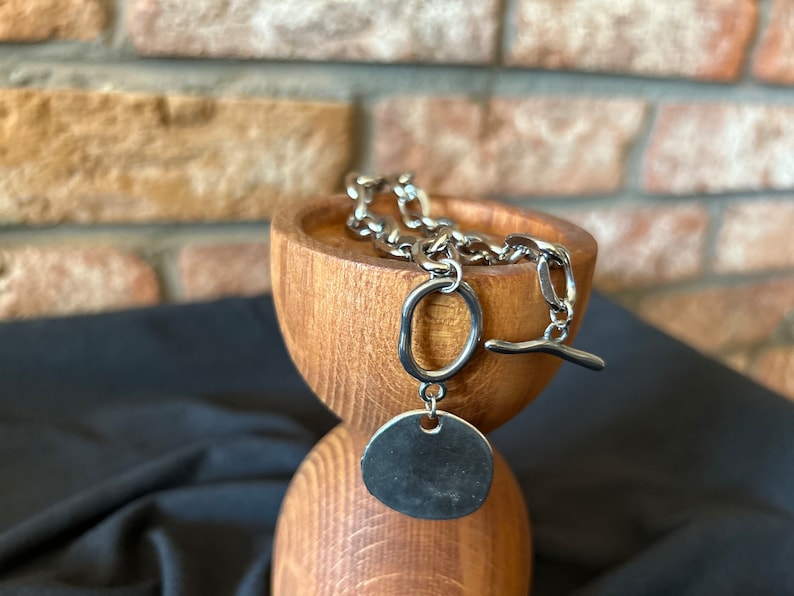 May include: A silver chain necklace with a large, round pendant. The chain is made of thick, linked metal and the pendant is smooth and shiny. The necklace is displayed on a wooden stand.