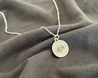 May include: A silver-colored necklace with a circular pendant featuring an eye design. The pendant is attached to a delicate chain. The necklace is displayed on a dark gray fabric surface.