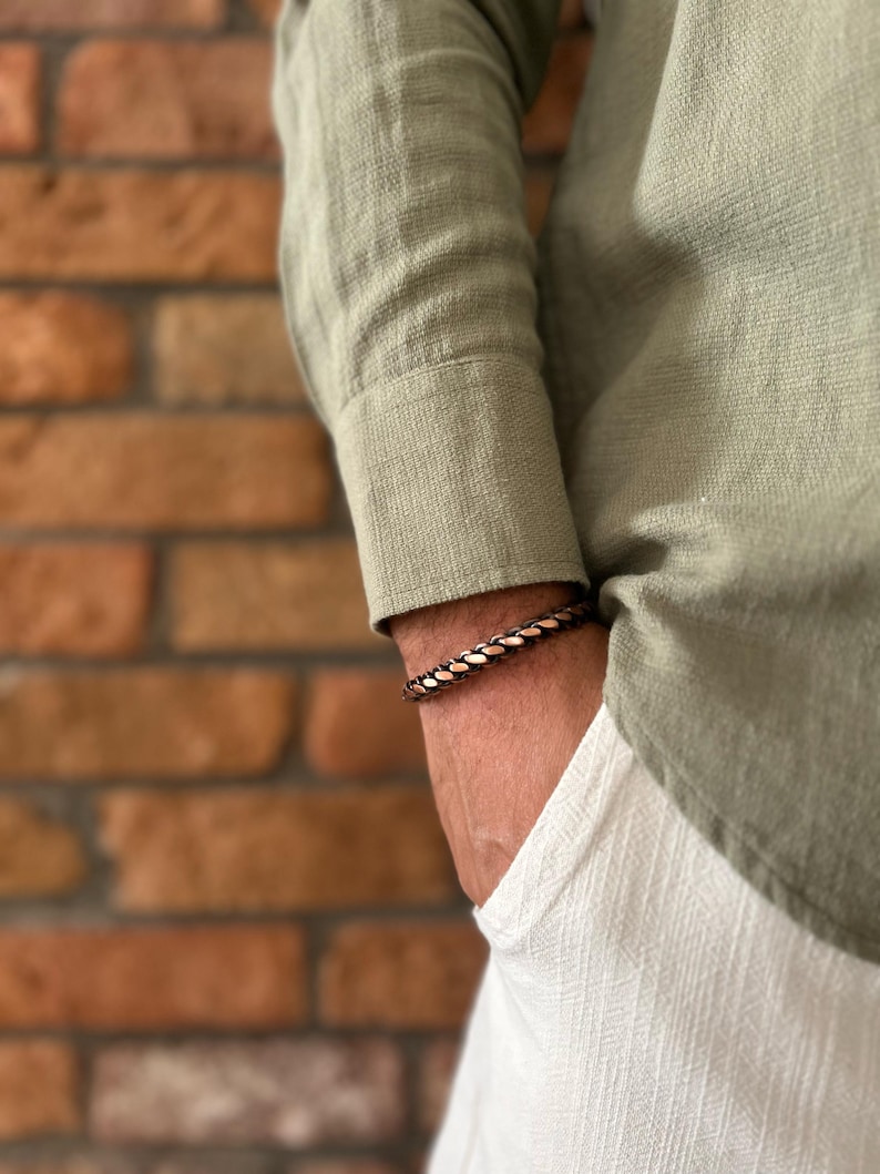 K&ouml;nnte beinhalten: Ein kupferfarbenes geflochtenes Armband wird am Handgelenk getragen, mit einem gr&uuml;nen Langarmhemd und einer wei&szlig;en Hose im Hintergrund. Das Armband besteht aus ineinander verwobenem Kupfer und bronzefarbenem Metall.