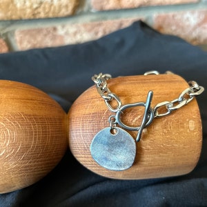 May include: A silver chain bracelet with a large, round, silver charm. The bracelet has a toggle clasp.