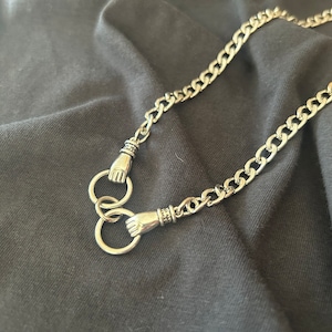 May include: A silver chain necklace with two hand-shaped clasps. The chain is thick and has a curb link design.