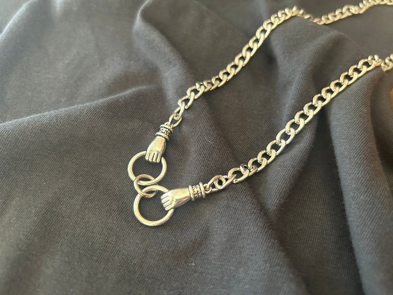May include: A silver chain necklace with two hand-shaped clasps. The chain is thick and has a curb link design.