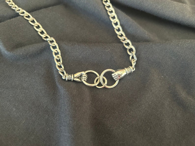 May include: A silver chain necklace with two linked rings, each ring has a hand-shaped design.