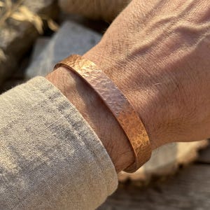 Può includere: Un bracciale a fascia in rame caratterizzato da una superficie martellata e testurizzata. La banda metallica presenta una calda finitura color bruno-rossastro e una larghezza costante che avvolge il polso. Il design è semplice e lineare.
