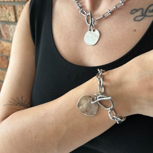 May include: A silver chain necklace and bracelet with large, round, silver pendant charms. The necklace and bracelet have a chunky, link chain design.