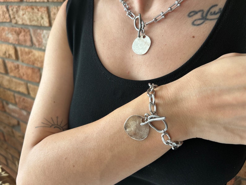 May include: A silver chain necklace and bracelet with large, round, silver pendant charms. The necklace and bracelet have a chunky, link chain design.