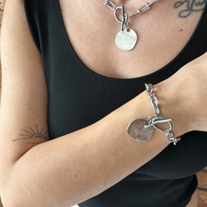 May include: A silver chain necklace with a large, round pendant and a matching silver chain bracelet with a similar pendant. Both pieces of jewelry feature a chunky, link chain design.