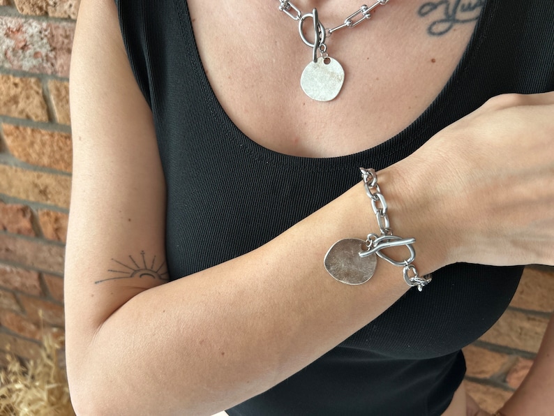 May include: A silver chain necklace with a large, round pendant and a matching silver chain bracelet with a similar pendant. Both pieces of jewelry feature a chunky, link chain design.