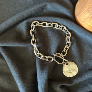 May include: A silver chain necklace with a large, round pendant. The chain is made of thick, chunky links. The pendant is smooth and has a slightly textured surface.