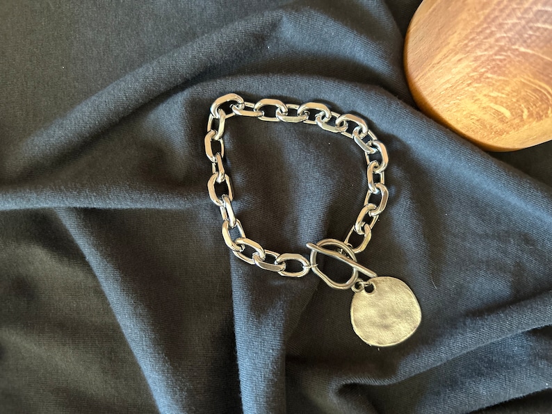 May include: A silver chain necklace with a large, round pendant. The chain is made of thick, chunky links. The pendant is smooth and has a slightly textured surface.