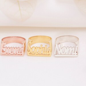 Puede incluir: Tres anillos de nombre personalizados con los nombres "Emma", "Sophia" y "Nomi" en una fuente cursiva. Los anillos están hechos de diferentes metales: oro rosa, oro amarillo y plata.
