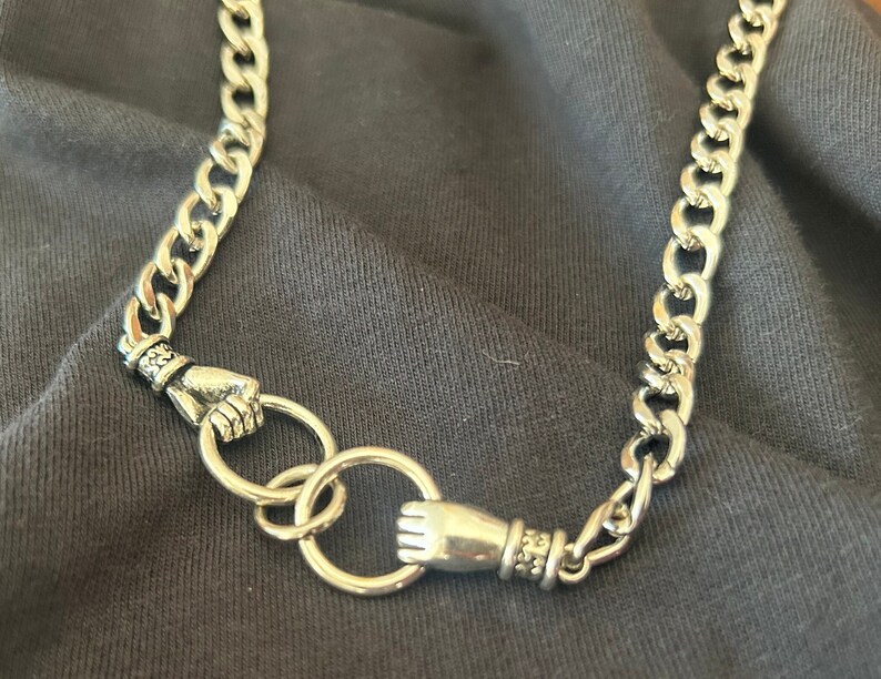 May include: A silver chain necklace with two silver hand-shaped clasps. The chain is a curb style.