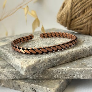 K&ouml;nnte beinhalten: Ein kupferfarbenes Armband mit kettenartigem Design. Das Armband ist offen und liegt auf einem Stapel grauer Steinplatten. Das Kupfer hat einen warmen, r&ouml;tlich-braunen Farbton, und die Glieder sind leicht l&auml;nglich.