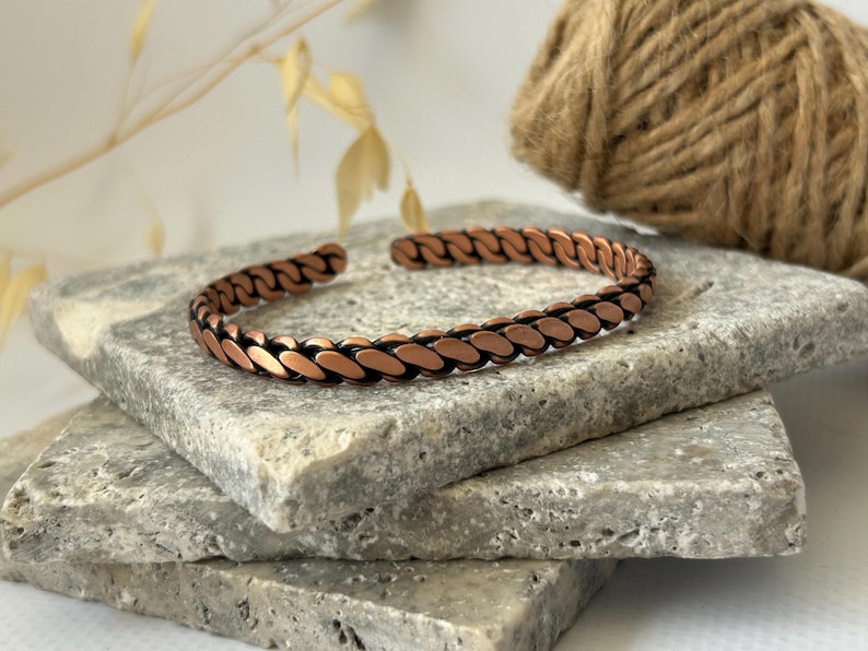 K&ouml;nnte beinhalten: Ein kupferfarbenes Armband mit kettenartigem Design. Das Armband ist offen und liegt auf einem Stapel grauer Steinplatten. Das Kupfer hat einen warmen, r&ouml;tlich-braunen Farbton, und die Glieder sind leicht l&auml;nglich.
