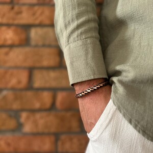K&ouml;nnte beinhalten: Ein geflochtenes Kupferarmband in Braun- und Goldt&ouml;nen wird am Handgelenk getragen. Das Armband befindet sich an einer Person, die ein hellgr&uuml;nes Langarmhemd und eine wei&szlig;e Hose tr&auml;gt. Der Hintergrund ist eine Backsteinmauer.