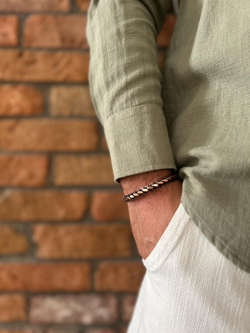 K&ouml;nnte beinhalten: Ein geflochtenes Kupferarmband in Braun- und Goldt&ouml;nen wird am Handgelenk getragen. Das Armband befindet sich an einer Person, die ein hellgr&uuml;nes Langarmhemd und eine wei&szlig;e Hose tr&auml;gt. Der Hintergrund ist eine Backsteinmauer.