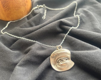 May include: A silver chain necklace with a silver pendant shaped like an eye. The eye is closed and has a detailed design.