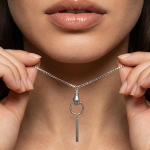 May include: Silver-toned necklace featuring a pendant with a hand holding a ring and a vertical bar. The necklace is held up, showcasing the design. The chain is delicate and the pendant is detailed.