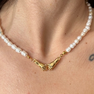 Op de afbeelding: Een witte parelketting met een gouden hanger met twee handen die in elkaar grijpen.