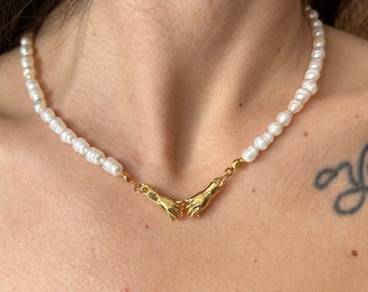 May include: A white pearl necklace with a gold pendant featuring two hands clasped together.
