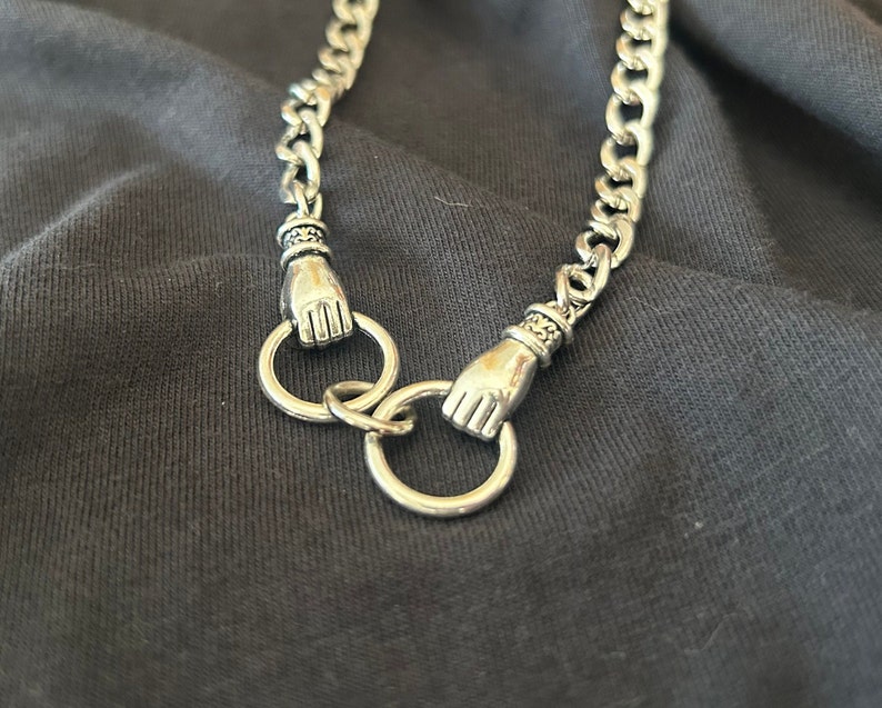 May include: A silver chain necklace with two hand-shaped clasps. The chain is thick and has a curb link design.