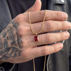 May include: A gold-toned chain necklace featuring a rectangular, faceted deep red gemstone pendant held by four prongs. The pendant hangs from a thin, smooth metal chain, showcasing a classic design with a vibrant, polished red stone center.