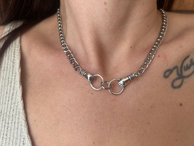 May include: A silver chain necklace with two hand-shaped clasps connected by two silver rings.