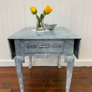 Puede incluir: Una pequeña mesa auxiliar gris y desgastada con un cajón y patas torneadas. La mesa tiene un acabado blanqueado y un diseño floral decorativo en la parte delantera del cajón. Dos rosas amarillas están en un jarrón de vidrio sobre la mesa.