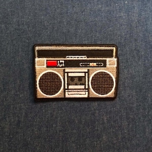 May include: An embroidered patch shaped like a retro boombox in shades of grey, black, and white. The design features two circular speakers, a central cassette deck compartment, and a small red rectangular accent on the upper control panel.
