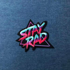 May include: Embroidered patch with the words "STAY RAD" in a vibrant gradient of teal, pink, and purple. The text is outlined in white and set against a black, star-shaped background. The patch is on a blue denim surface.