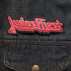 May include: A close-up shot of a dark denim jacket with a red and black embroidered patch. The patch displays the band name "Judas Priest" in a stylized font. A pocket with a button is visible below the patch.
