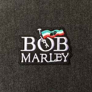 May include: Embroidered patch featuring the name "BOB MARLEY" in white block letters on a black background. A small flag with red, white, green, and black stripes is above the "B". The patch is on a dark gray fabric.