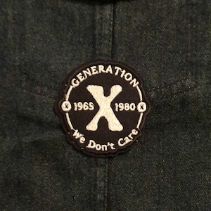 May include: Dark denim jacket with a circular patch. The patch features the text "GENERATION X" around a white "X" with the years 1965 and 1980. The phrase "We Don't Care" is also on the patch. A brass button is visible.