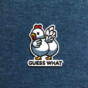 May include: Embroidered patch of a cartoon chicken with a white body, blue detailing, and a red comb. The patch includes the text "GUESS WHAT" in a bold, black font, outlined in white. The patch is set against a blue denim background.