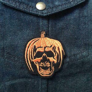 May include: Embroidered patch with an orange and black jack-o'-lantern skull design. The patch is attached to dark blue denim fabric, near a pocket with a silver button. A Halloween-themed accessory, suitable for clothing.