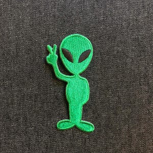 May include: Green embroidered alien patch with black eyes and a peace sign hand gesture on a dark denim background. The alien is a classic cartoon style.