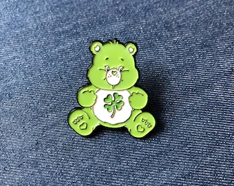 May include: A green Care Bear enamel pin with a black outline. The bear is sitting and has a white belly with a four-leaf clover design. The pin is on a blue denim background.