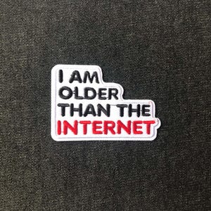 May include: An embroidered patch with the text "I AM OLDER THAN THE INTERNET." The patch features white, black, and red colours, with a white border. It is set against a dark grey textured background.
