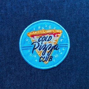 May include: A circular embroidered patch on a dark blue denim fabric. The patch features a light blue background with a central graphic of a pizza slice in yellow and red. The text COLD PIZZA CLUB is embroidered in dark blue across the slice.