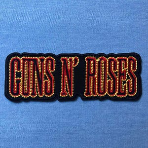 May include: Embroidered patch featuring the text "GUNS N' ROSES" in red, outlined with gold beads, on a black background. The patch has a black border and is set against a light blue fabric.