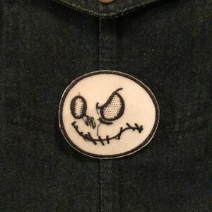 K&ouml;nnte beinhalten: Ein ovales Patch mit einem schwarz-wei&szlig; gestickten Jack Skellington-Gesicht. Das Patch ist auf einer dunklen Jeansjacke befestigt. Das Design umfasst gestickte Augen, einen gestickten Mund und eine gestickte Nase. Die Jacke hat einen Messingknopf an der Tasche.