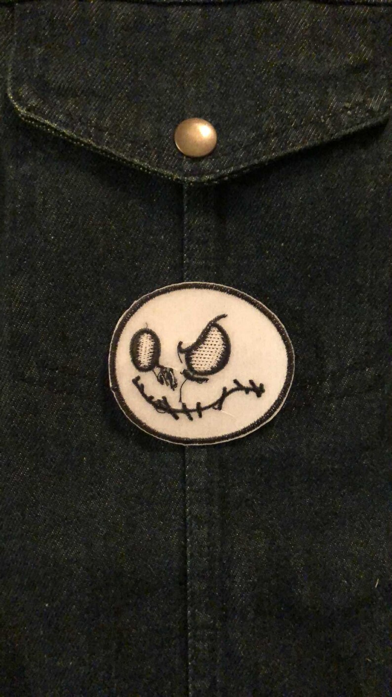 K&ouml;nnte beinhalten: Ein ovales Patch mit einem schwarz-wei&szlig; gestickten Jack Skellington-Gesicht. Das Patch ist auf einer dunklen Jeansjacke befestigt. Das Design umfasst gestickte Augen, einen gestickten Mund und eine gestickte Nase. Die Jacke hat einen Messingknopf an der Tasche.