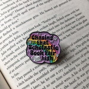 May include: A pin with the text "Chasing that Scholastic Book Fair high" in a cloud shape with a rainbow and flowers. The pin is on a book page.