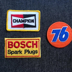 May include: Three embroidered patches: "Champion" in black and red, "BOSCH Spark Plugs" in red and yellow, and a round orange patch with "76" in blue. All patches are on a dark blue denim background.