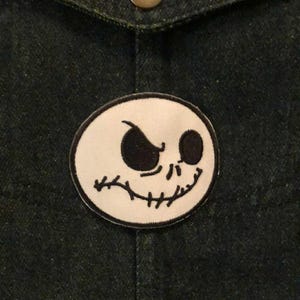K&ouml;nnte beinhalten: Dunkle Jeansjacke mit einem runden Aufn&auml;her mit Jack Skellingtons Gesicht aus The Nightmare Before Christmas. Der Aufn&auml;her ist wei&szlig; mit schwarzem Umriss und schwarzen Gesichtsz&uuml;gen. Ein Messingknopf ist sichtbar.