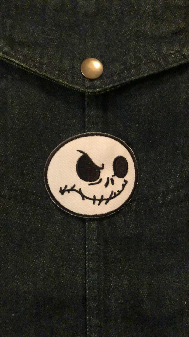 K&ouml;nnte beinhalten: Dunkle Jeansjacke mit einem runden Aufn&auml;her mit Jack Skellingtons Gesicht aus The Nightmare Before Christmas. Der Aufn&auml;her ist wei&szlig; mit schwarzem Umriss und schwarzen Gesichtsz&uuml;gen. Ein Messingknopf ist sichtbar.