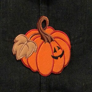 Peut inclure: Applique de citrouille orange avec un visage souriant, une tige et une feuille brunes, fixée sur une veste en jean bleu foncé. La citrouille est un article sur le thème de l'automne ou d'Halloween.